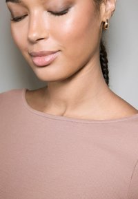 Close-up of a model's upper body wearing a light pink ribbed top with a wide neckline, featuring smooth skin and small gold hoop earrings.