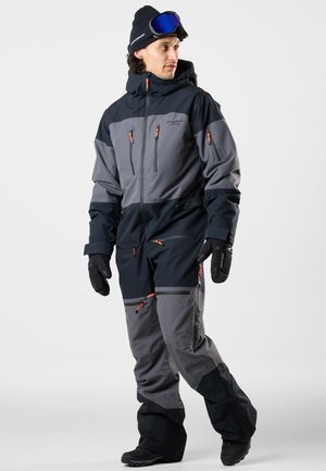 Mand iført sort og grå skidragt med orange lynlåse, sorte handsker, sort hue og blå skibriller, der kigger til siden.