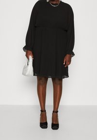 Robe plissée noire avec des manches longues, un col rond et une coupe décontractée. Les accessoires incluent un sac à main en perles argentées et des talons noirs à bride cheville.