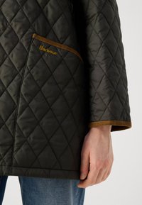 Gequilted donkergroen jack met bruine accenten, voorzien van een borstzak en geribbelde manchetten. Merkennaam "Barbour" geborduurd op de zak.