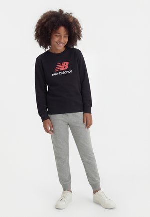 Bambino con capelli ricci che indossa una felpa nera New Balance, pantaloni della tuta grigi e scarpe da ginnastica bianche, in piedi e sorridente contro uno sfondo bianco.