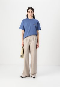 Blaues Baumwoll-T-Shirt mit einem kleinen gestickten Logo, kombiniert mit hochgeschnittenen beigen Hosen und einer hellbeigen Tasche mit Griff.