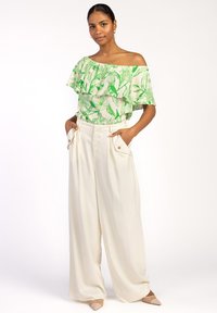 Off-shoulder top met groen bladerenpatroon op een lichte achtergrond, gecombineerd met hooggepolsterde crèmekleurige broek met wijde pijpen en zijzakken.