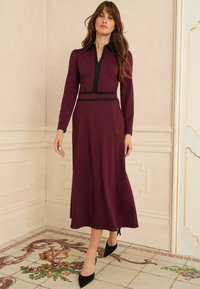 Rochie lungă de culoare maroon, cu un guler și un brâu negru cu fermoar, având mâneci lungi și o fustă vaporoasă. Pur și simplu cu tocuri negre ascuțite.