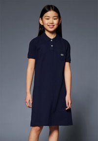 Rochie polo bleumarin din bumbac, cu guler, mâneci scurte și un buzunar pe piept. Prezintă un logo verde pe buzunar. Croială simplă și dreptă.