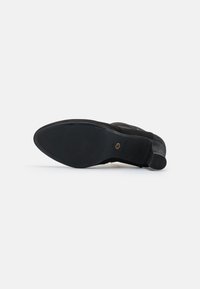 Zapato de ante negro con suela de goma, que presenta un talón texturizado y un diseño suave y aerodinámico. Detalle del logo visible cerca del talón.