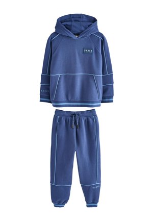 Adolescente in piedi che indossa una felpa con cappuccio blu abbinata e pantaloni della tuta con sneakers bianche su uno sfondo semplice.