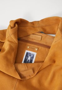 Brown suede jacket interior with a zipper pocket, branded label, and accent stitching. Soft texture, featuring a folded collar.