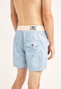 Bañadores para hombre en azul claro con un patrón geométrico, cinturilla elástica con un acento en crema y un bolsillo trasero con un botón de madera.