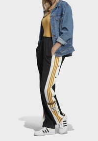 Person som bär svarta träningsbyxor med gula ränder och tryckknappar, vita Adidas sneakers, en gul topp och en blå denimjacka.