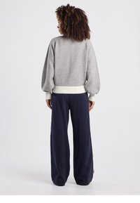 Pull gris à manches bouffantes avec un ourlet côtelé crème, associé à un pantalon large bleu marine, le tout dans un décor neutre.