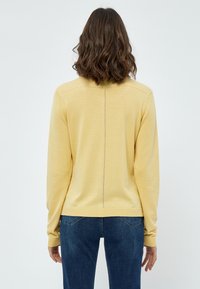 Gul stickad tröja med långa ärmar, strukturerat mönster längs ryggen och ribbad mudd och nederkant. Matchas med blå jeans.