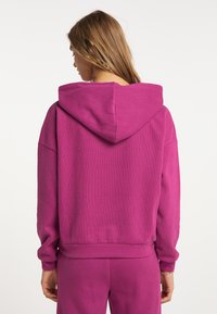 Felpa viola con cappuccio, texture a coste, spalle scese e orlo dritto, indossata sopra pantaloni abbinati, che mostra un look casual.