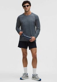 Športový outfit pozostávajúci z tmavosivej dlhého rukávu s textúrovaným dizajnom, čiernych šortiek, sivých tenisiek a fľaše na vodu.