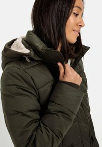 Olivengrøn pufferjakke med hætte i faux fur og ribbede manchetter. Har en quiltet tekstur og lynlås foran.