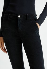 Pantaloni in corduroy neri con una texture liscia, una tasca frontale e un bottone argentato in vita, abbinati a una maglietta a maniche lunghe a coste in blu navy.