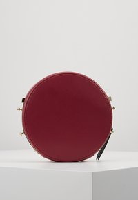 Borsa tonda in pelle bordeaux con texture liscia, dettagli in metallo dorato e chiusura a zip. Design semplice e minimalista.