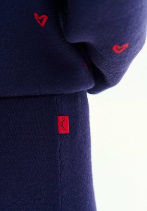 Granatowa dzianina z małymi czerwonymi haftowanymi sercami oraz czerwona metka z symbolem czarnej rzęsy na szwie.