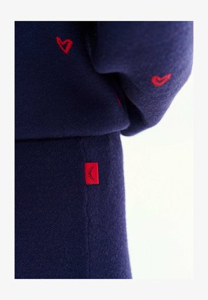 Granatowa dzianina z małymi czerwonymi haftowanymi sercami oraz czerwona metka z symbolem czarnej rzęsy na szwie.