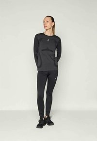 Vrouw in zwarte sportieve lange mouw top en leggings met logo's, staand met handen achter de rug, kijkend opzij tegen een effen achtergrond.