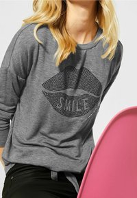 Graues Langarmshirt mit einem gepunkteten Lippen-Graphic und dem Wort "SMILE" sowie einer weichen Textur. Dargestellt vor einem rosa Stuhl.