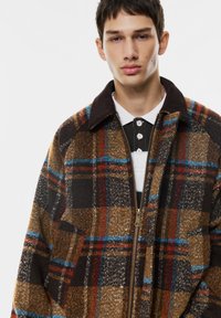 Hnedo-čierny károvaný kabát s modrými a červenými akcentmi, s predným zipsom, širokým golierom a štruktúrovanou textíliou.