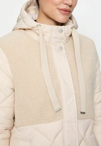 Frau, die eine cremefarbene gesteppte Jacke mit einem flauschigen Einsatz auf der Brust und einer Kapuze mit cremefarbenen Kordeln trägt.