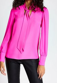 Blusa rosa brillante con maniche lunghe e scollatura annodata. Tessuto liscio, con apertura a chiave sul davanti e polsini abbottonati.
