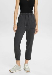 Femme portant un pantalon tapered charbon avec des poches avant à rabat et des baskets gris clair, montrée des épaules aux pieds.