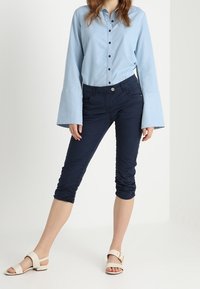 Kvinna klädd i en ljusblå långärmad skjorta med utsvängda manschetter, marinblå korta ruched byxor och vita sandaler med spännen.