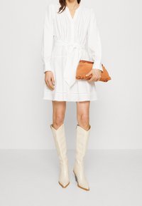 Vit klänning med långa puffärmar, midjesnörning och ögonletsdetaljer. Sammanställd med knähöga krämfärgade stövlar och en orange kuvertväska.