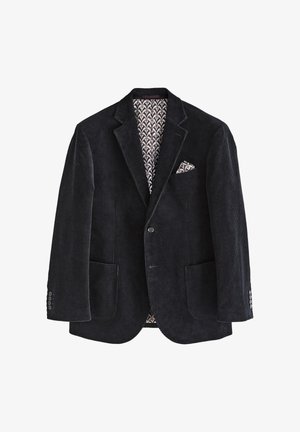 Man draagt een donker corduroy colbert met pochet, zwarte trui, wit overhemd met kraag, zwarte broek en zwarte schoenen, kijkt opzij.