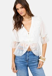 Blusa bianca trasparente con texture a pois, bordo in pizzo e scollatura arricciata. Abbinata a jeans blu a vita alta e bracciali impilati.
