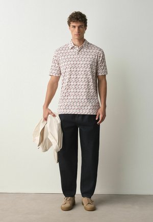 Junger Mann steht, trägt ein kurzärmeliges Poloshirt mit Muster, schwarze Hose, braune Schuhe und hält eine gefaltete beige Jacke in der linken Hand.