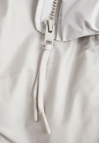 Světlá šedá látka bundy s detailním záběrem na zip, který je vybaven texturovaným táhlem a nastavitelnou šňůrkou s potaženými konci.
