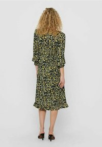 Vestido negro con patrón floral amarillo, de manga larga y con dobladillo con volantes. Hecho de una tela ligera, tiene una cintura ajustada y una falda acampanada.