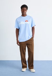 Gai zilbā kokvilnas T-krekls ar baltu "ellesse" logotipu un sarkanu akcentu, kombinēts ar brūnām brīvā griezuma biksēm un krēmkrāsas kedām.