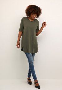 Tunique olive verte avec un col rond, des manches courtes et des fentes sur les côtés, associée à un jean bleu ajusté et des ballerines noires à bout pointu.