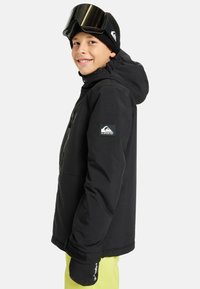 Uśmiechnięte dziecko w czarnej zimowej kurtce, czapce typu beanie, i goglach narciarskich, w czarnych rękawiczkach i jasnooliwkowych spodniach, stojące bokiem na białym tle.