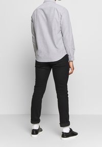 Svetle šedá košile na knoflíky s dlouhými rukávy, zaobleným dolním lemem, spárovaná s černými slim fit džíny a černými teniskami, předvádějící ohrnuté rukávy a bílé ponožky.