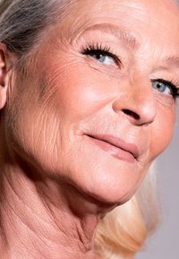 Närbild av ett ansikte med ljus hy, grått hår, slät textur, fina rynkor, ljusblå ögon och naturlig sminkning med nude läppstift.