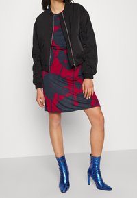 Veste bomber noire, robe ajustée fleurie bleu marine et bordeaux, bottes à talons brillantes bleues. Détails en métal zippé et tissu texturé.