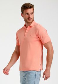 Kortärmad polo t-shirt i ljus korallfärg, med klassisk krage, knapplist och diskret logotyp på bröstet. Slät bomullstextur.