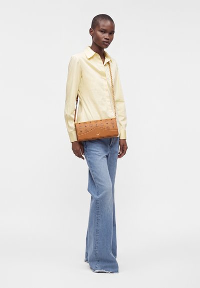 Femme portant une chemise jaune pâle et un jean bleu à jambes larges, debout avec un sac bandoulière en cuir beige avec une sangle chaîne dorée.