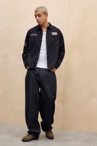 Fekete, gombos ing fehér hímzett részletekkel, laza sötét farmernadrággal és barna cipővel párosítva. Lágy, sima anyagtextúrák jól láthatóak.