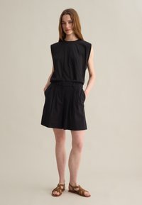 Camisola nera senza maniche con collo rotondo, abbinata a pantaloni corti neri a vita alta con una parte anteriore plissettata e tasche laterali. Sandali marroni con cinturini.