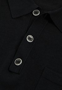 Polo cu un guler negru, cu o textură tricotată, având trei butoane argintii la guler și un mic buzunar pe piept.