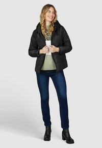 Schwarze Puffjacke mit Kapuze, Reißverschluss vorne und elastischen Bündchen, getragen über einem grünen Pullover und blauen Jeans, kombiniert mit schwarzen Blockabsatzstiefeln.