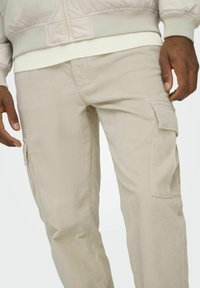 Beige kordfrigivna cargobyxor med en texturerad yta, som har sidofickor och en avslappnad passform, kombinerade med en ljus bomberjacka.