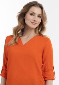 V-hals orange bluse med trekvartærmer, tekstureret stof og oprullede ærmer. Enkel design uden mønstre eller udsmykninger.